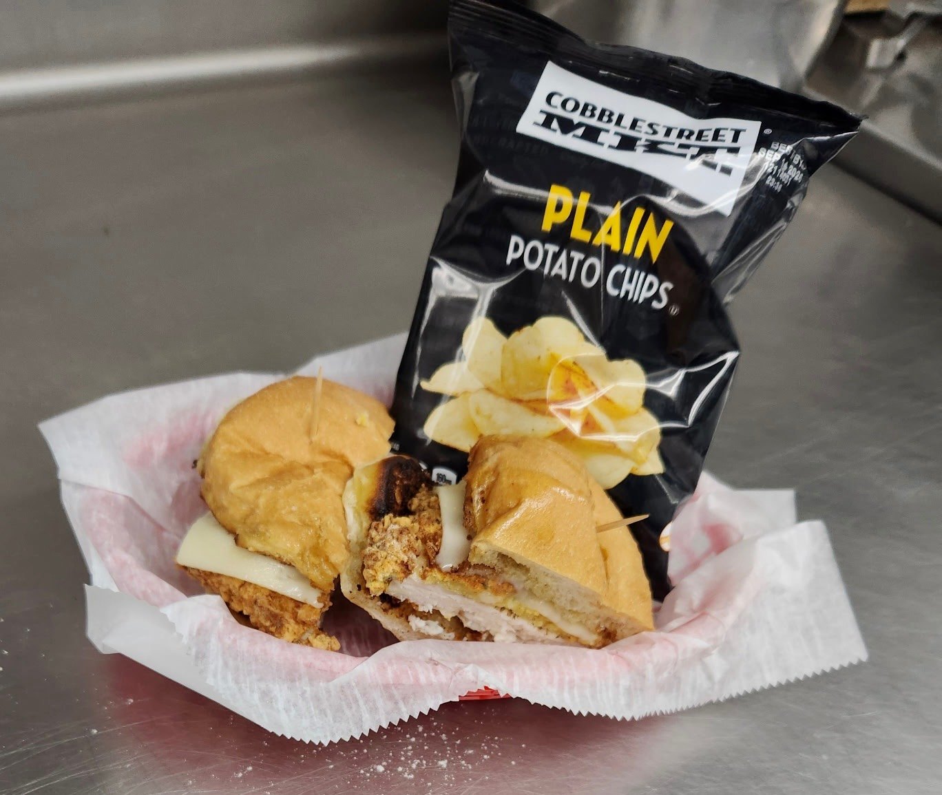 Crispy fried chicken sandwich cut in half with melted cheese, served with a bag of plain potato chips, from The Chicken Shack in Waupaca, WI